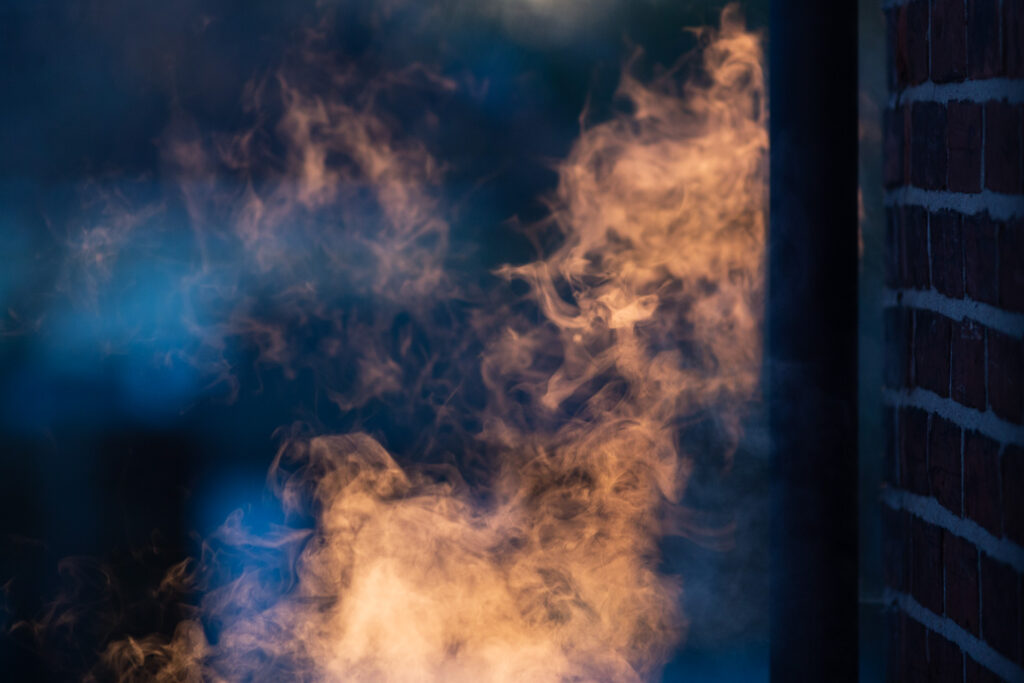 Photograph of steam that appears orange in the setting sun, against a dark background with a brick wall and drainpipe to the right.