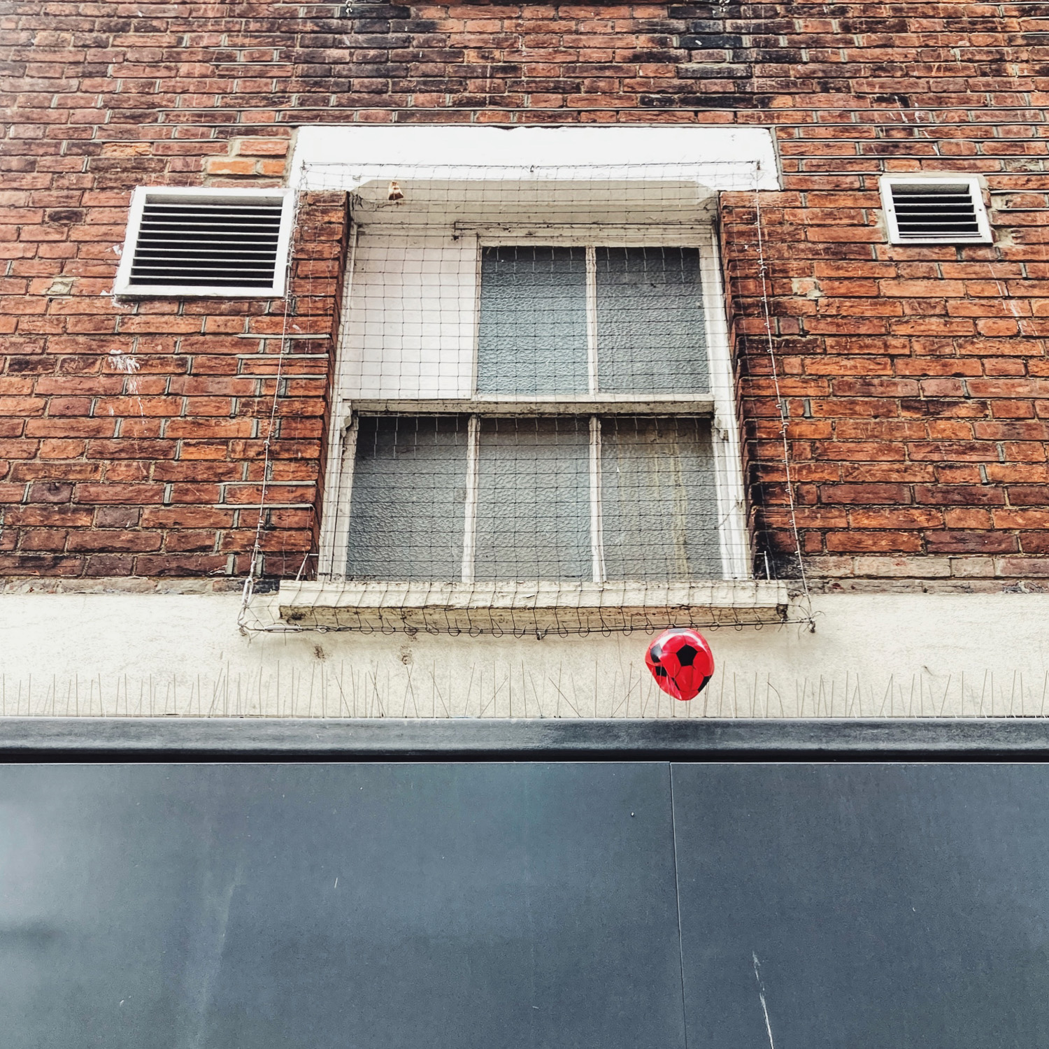 The end of a little red football. Photograph looking up at an old sash window in a brick wall, with air vents either side, a grey panel below which has spikes along it. A red inflatable football is stuck on one, deflating.