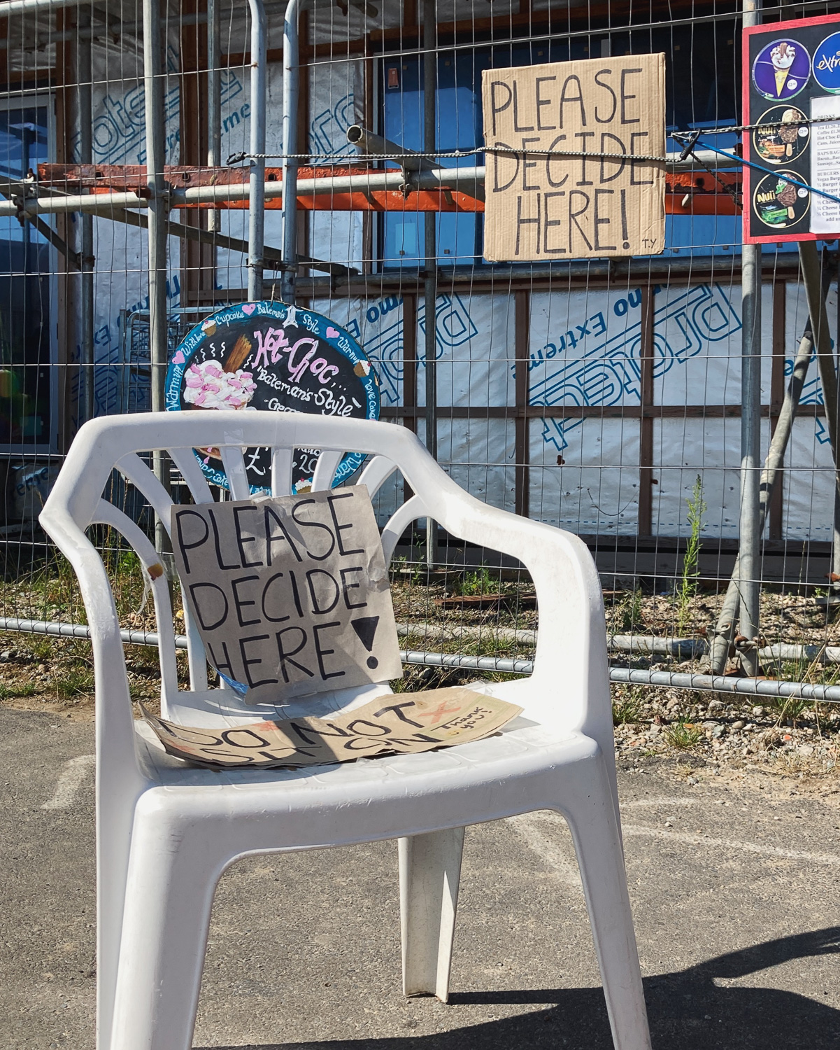 PLEASE DECIDE HERE! Photograph of a plastic garden chair with with a handwritten sign saying'PLEASE DECIDE HERE!', behind it is scafolding, another cardboard sign with the same message, and adverts for hot-choc and icecreams.