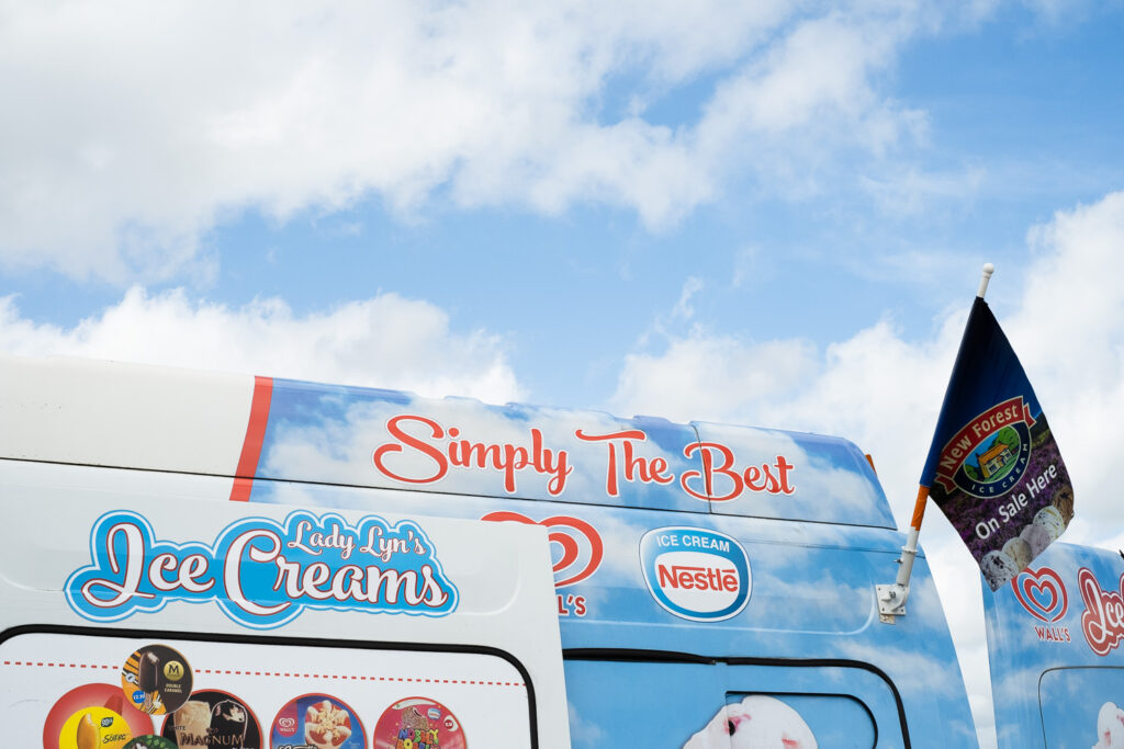 Photograph of a blue sky with clouds with the top of an ice cream van visible in the lower part of the frame, with a very similar sky scene painted on it, a flag saying 'New Forest Ice Cream On Sale Here' and text reading 'Lady Lyn's Ice Creams', 'Simply The Best' and 'Ice Cream Nestle' written on the van itself.