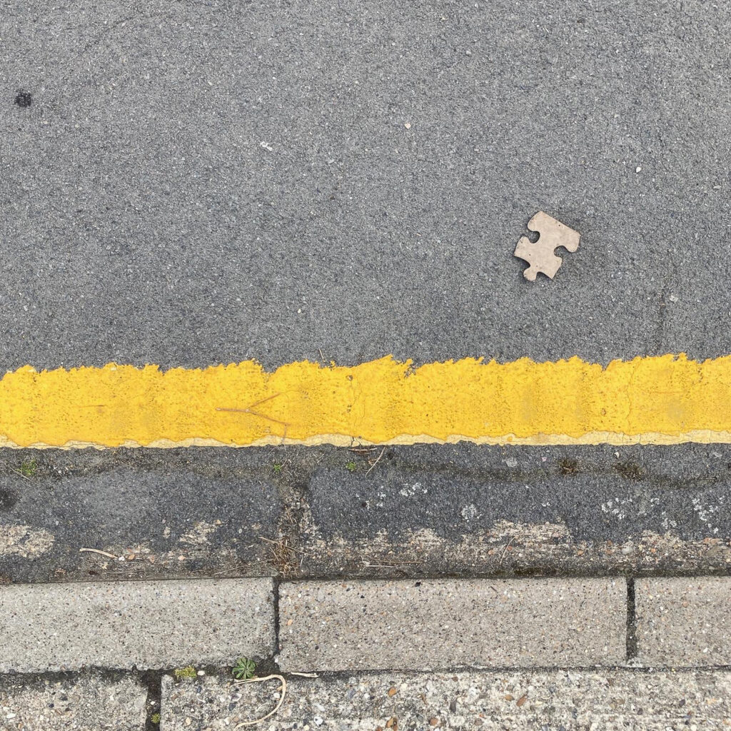 Photograph looking down at a gutter at the side of the road - a blank puzzle piece is in the top portion of the frame, with a single yellow line and pavement curb below.