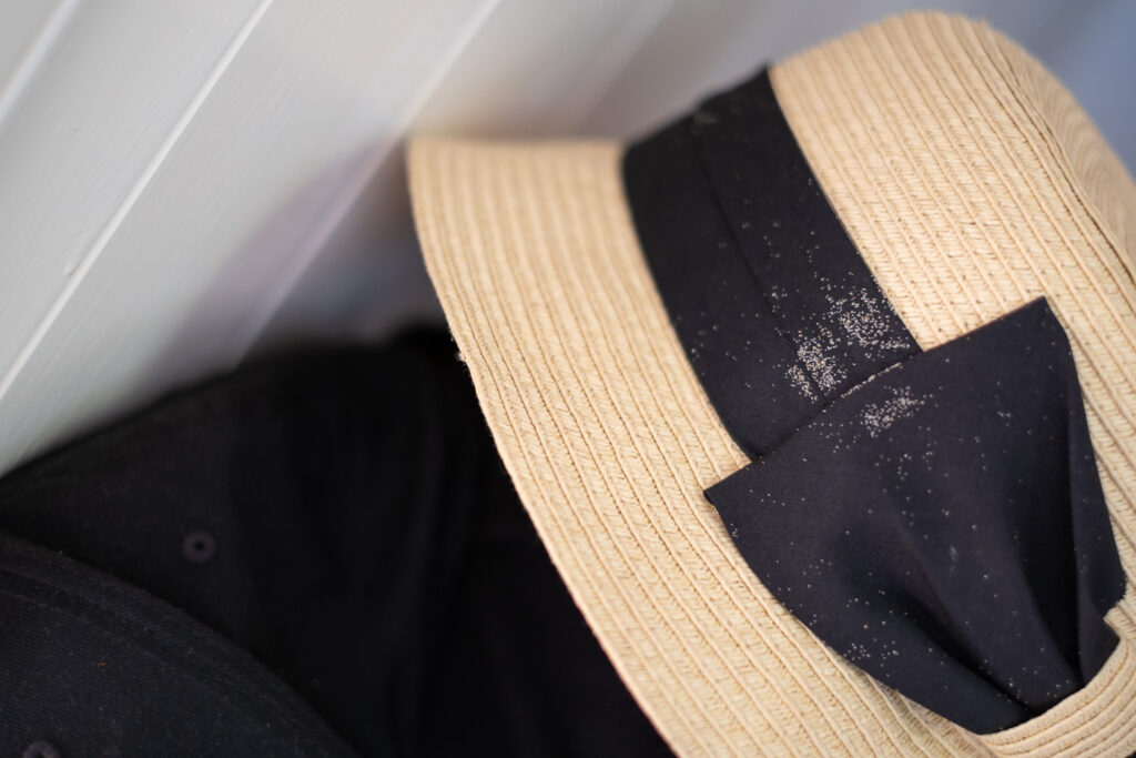 Photograph of a straw hat with grains of sand stuck to the black ribbon around the edge. The wooden underside of the stairs can be seen to the top left.