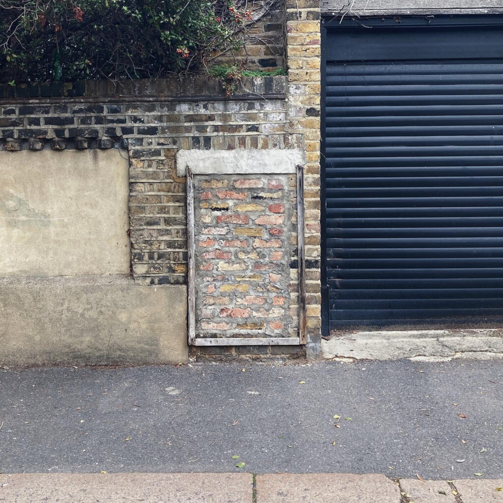 Photograph of a wall with the remains of a frame that used to hold a notice, with a black garage door to the right and pavement in front