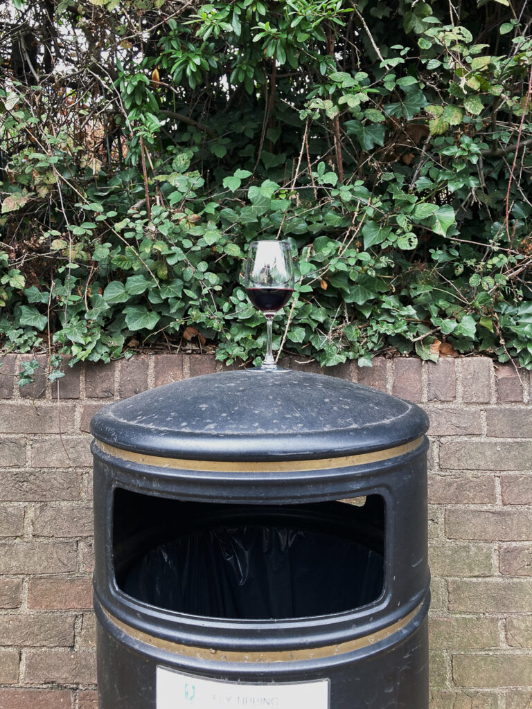 Photograph of a glass of red wine standing on top of a bin in a street, with a brick wall and foliage behind.