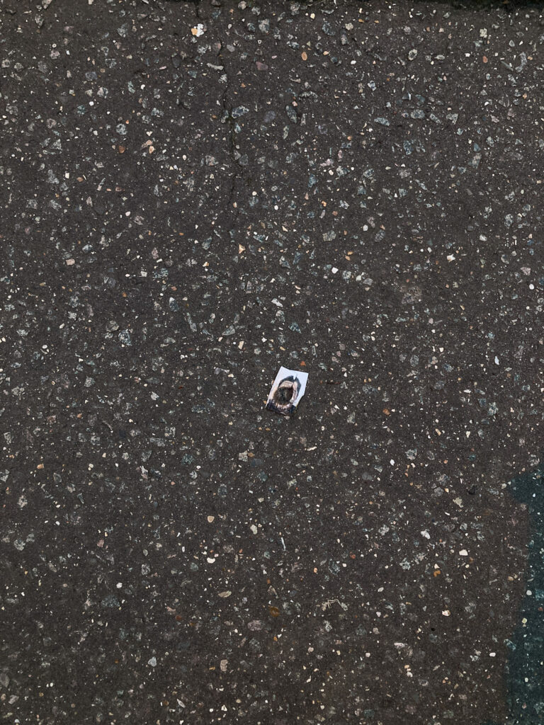 Photograph showing what looks like a passport photograph of a person, with the face burnt out to leave a circular hole, lying on a tarmace pavement.