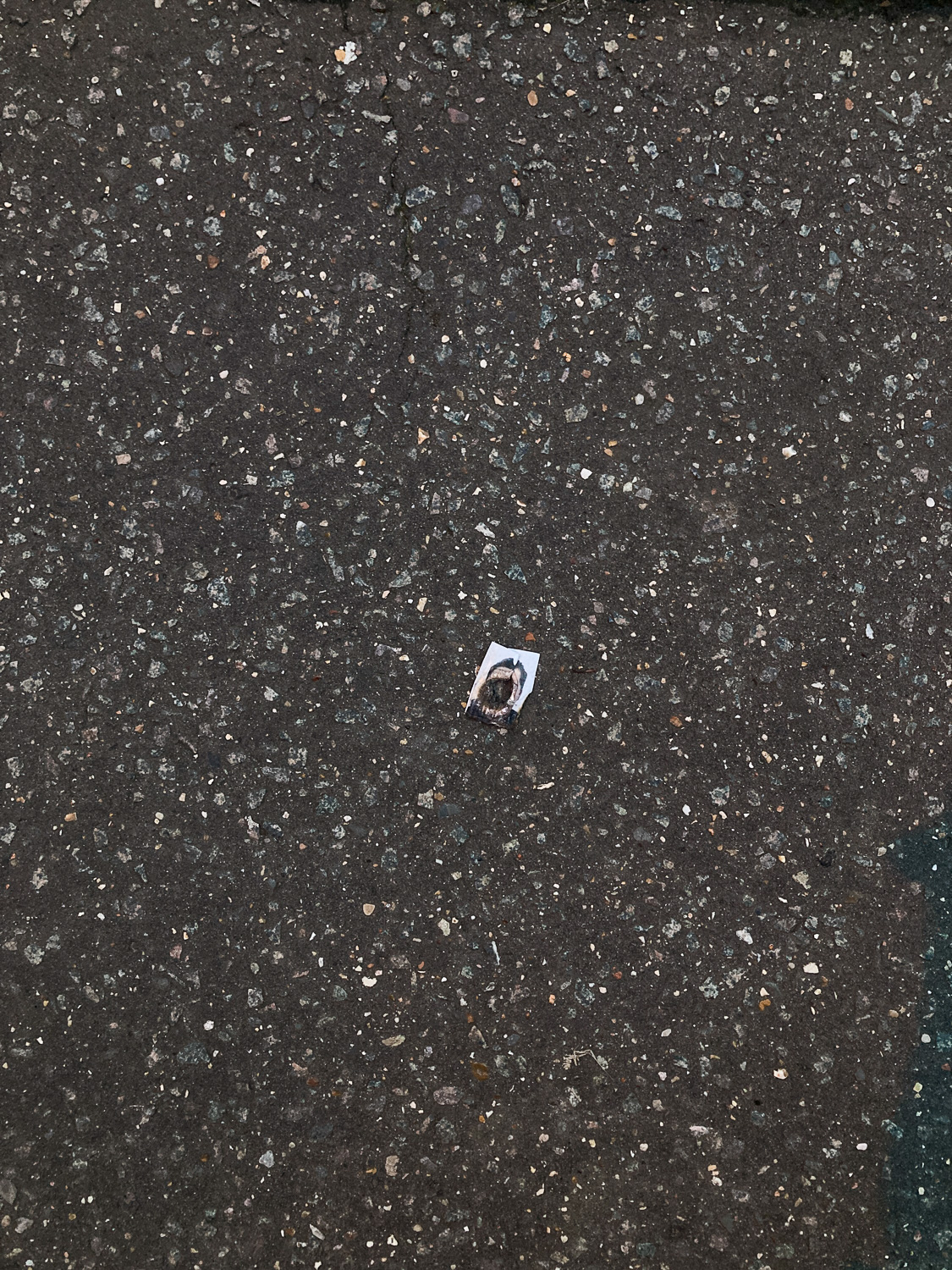 I hope they are okay. Photograph showing what looks like a passport photograph of a person, with the face burnt out to leave a circular hole, lying on a tarmace pavement.