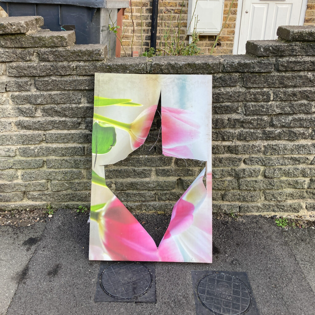 Photograph of a canvas artwork of flowers with a cross cut in the middle to make a big whole, leaning against a brick wall at the side of a pavement