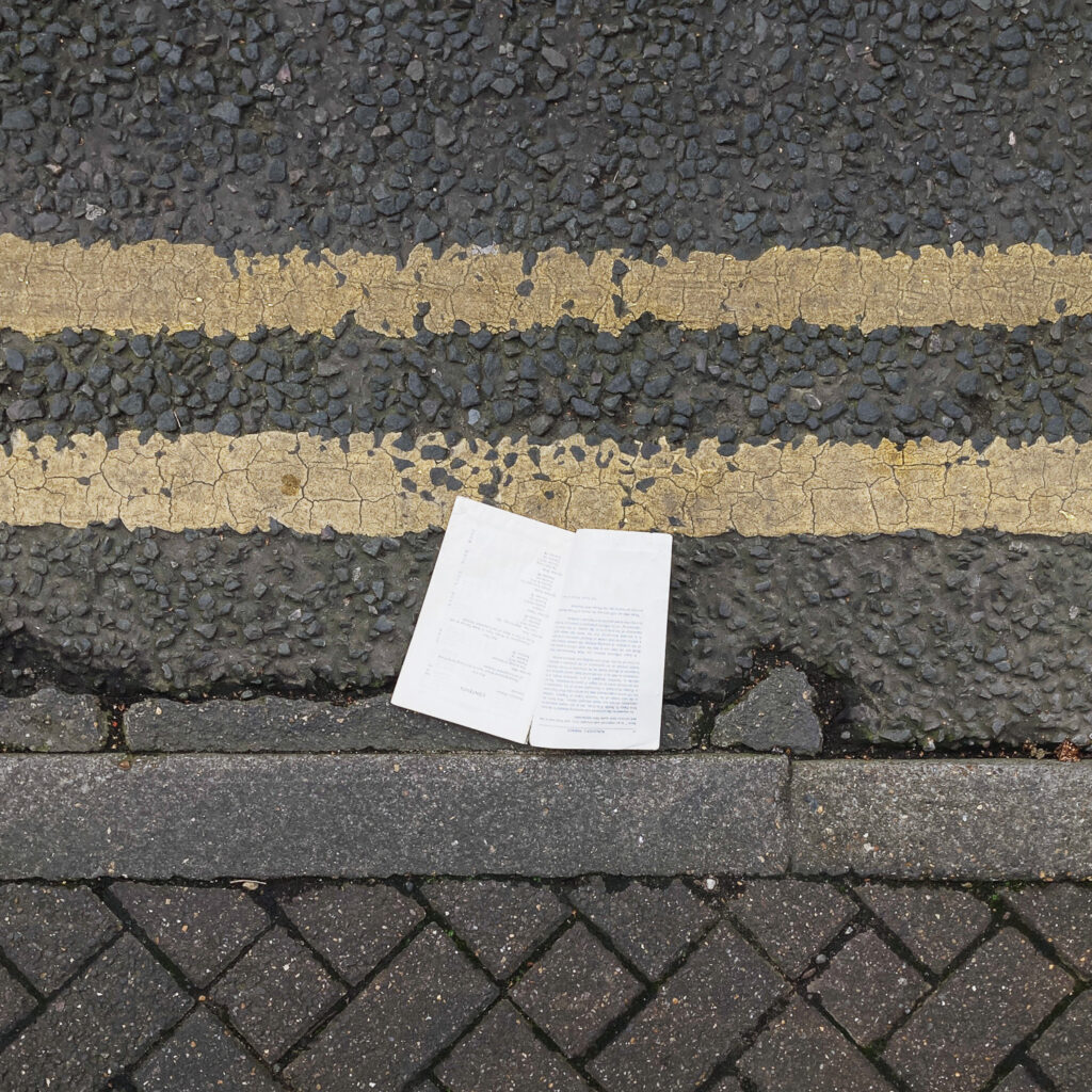 Photograph looking down at a gutter at the side of the road - an open portion of a paperback book is in the centre of the frame, with double yellow lines above and pavement curb below.