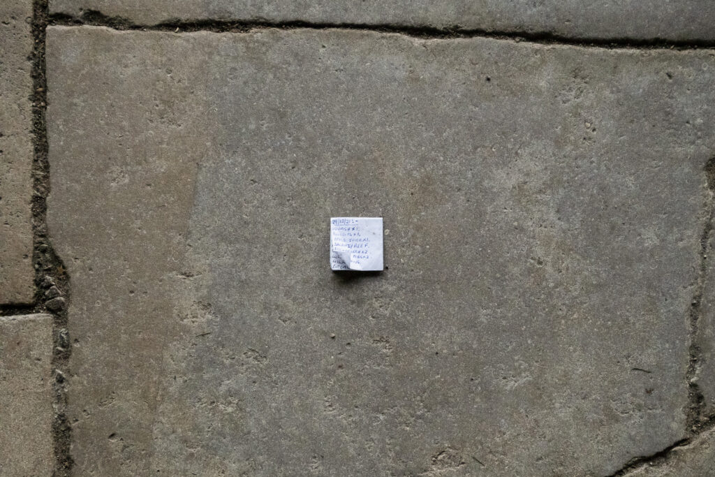 Photograph of a stone floor in Canterbury Cathedral with a small piece of paper with a handwritten shopping list that says '29/12/21:- SQUASH X 1. BISCUITS X 1. APPLE JUICE X 1. SALAMI/BEEF. DUO-CRUNCH X 2. ... PIES X 2'