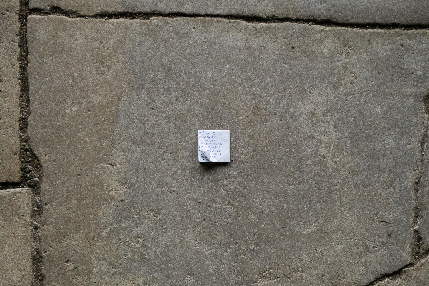 Photograph of a stone floor in Canterbury Cathedral with a small piece of pager with a handwritten shopping list that says '29/12/21:- SQUASH X 1. BISCUITS X 1. APPLE JUICE X 1. SALAMI/BEEF. DUO-CRUNCH X 2. ... PIES X 2 '
