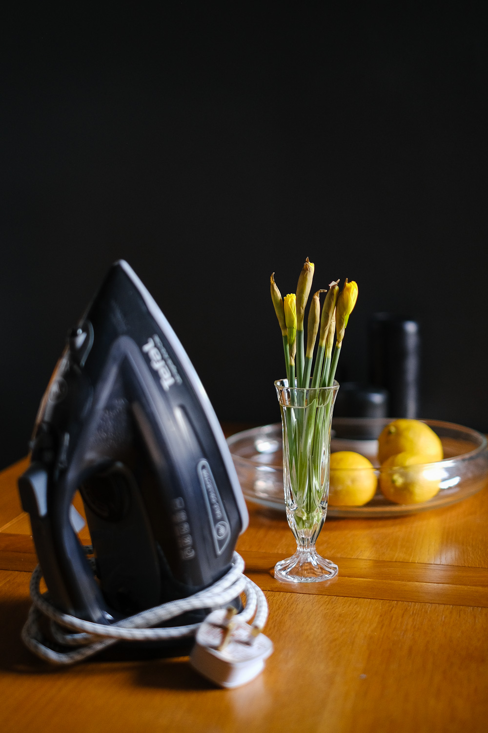 Still life with grey and yellow. Photograph of an iron, small glass vase of emerging daffodils, glass bowl with three lemons and black candles on a wooden table, with a dark grey wall behind.