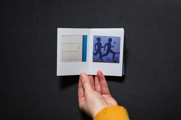 Photograph of hands holding a small foldy zine on a dark grey background. It is held open on pages with a photograph to the left of a blue handprint on a cream wall with a blue stripe to the right, and a photograph to the right of a mural showing dark purple painted runners on a lighter purple background, with black smeared handprints.