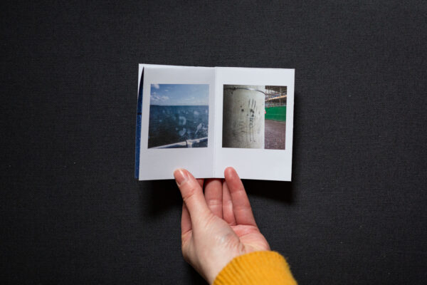 Photograph of hands holding a small foldy zine on a dark grey background. It is held open on pages with a photograph to the left of a white suncream handprint on a plastic screen with the sea and sky visible behind, and a photograph to the right of a concrete pillar with black smeared handprints.