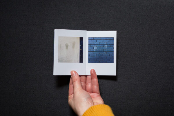 Photograph of hands holding a small foldy zine on a dark grey background. It is held open on pages with a photograph to the left of a blue handprint on a cream wall with three brown smeared handprints, and a photograph to the right of a black wall with 2 white smeared handprints.