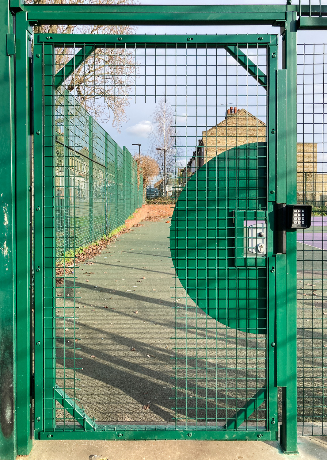 The introduction of a fee in a community tennis court. Photograph of a green metal gate with a lock and key pad to the right, a section of the gate has been cut away so people can still access the tennis court behind.