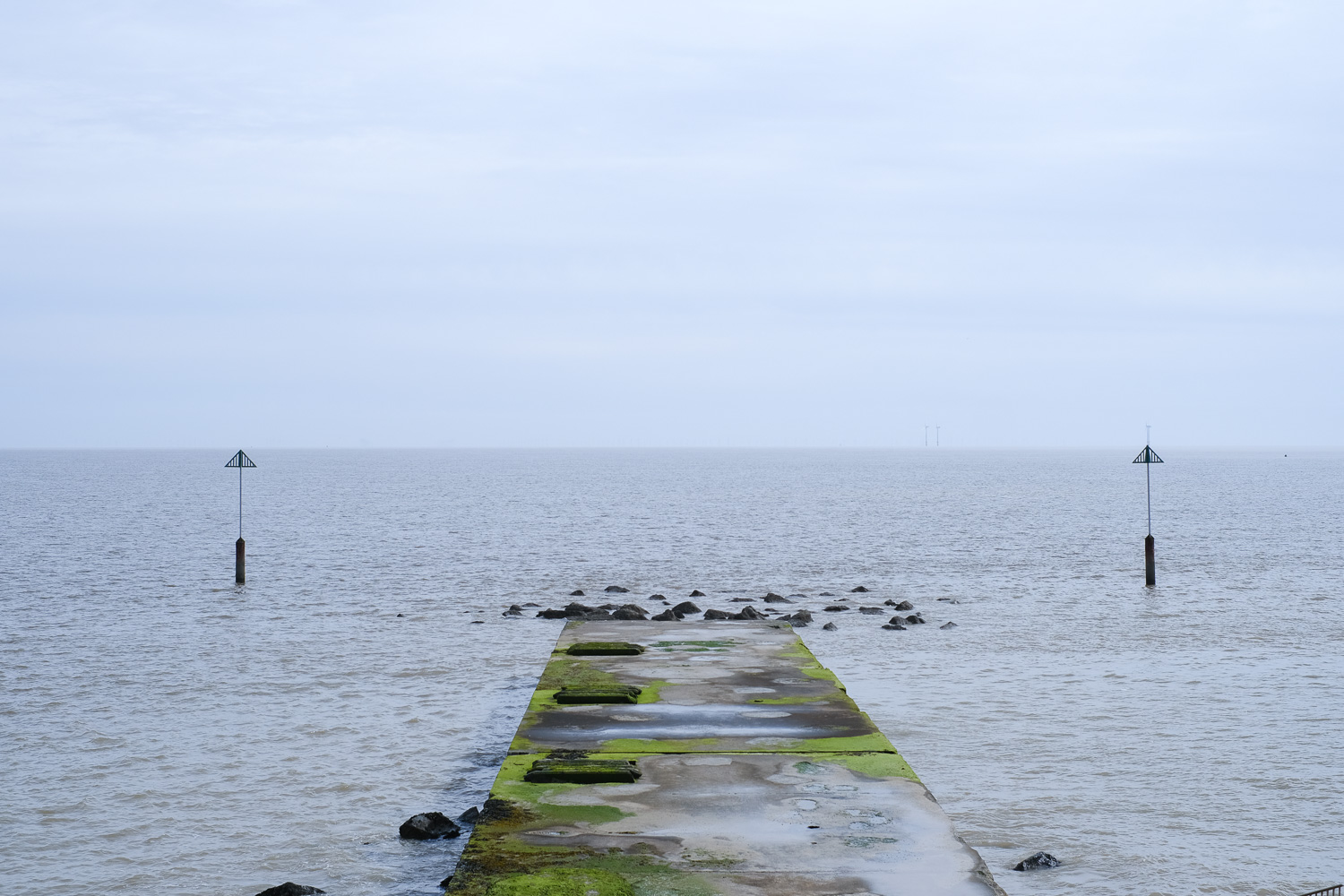 We escaped to the sea and it was wonderful. Photograph of a jetty covered in seaweed leading out into the sea, with two posts with triangles at the top to the left and right, and a pale blue sky above