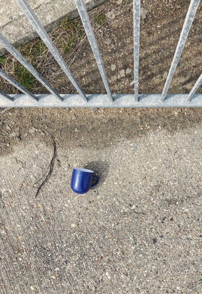 Photograph looking down at a blue mug on a concrete path, with metal railings at the top of the frame.