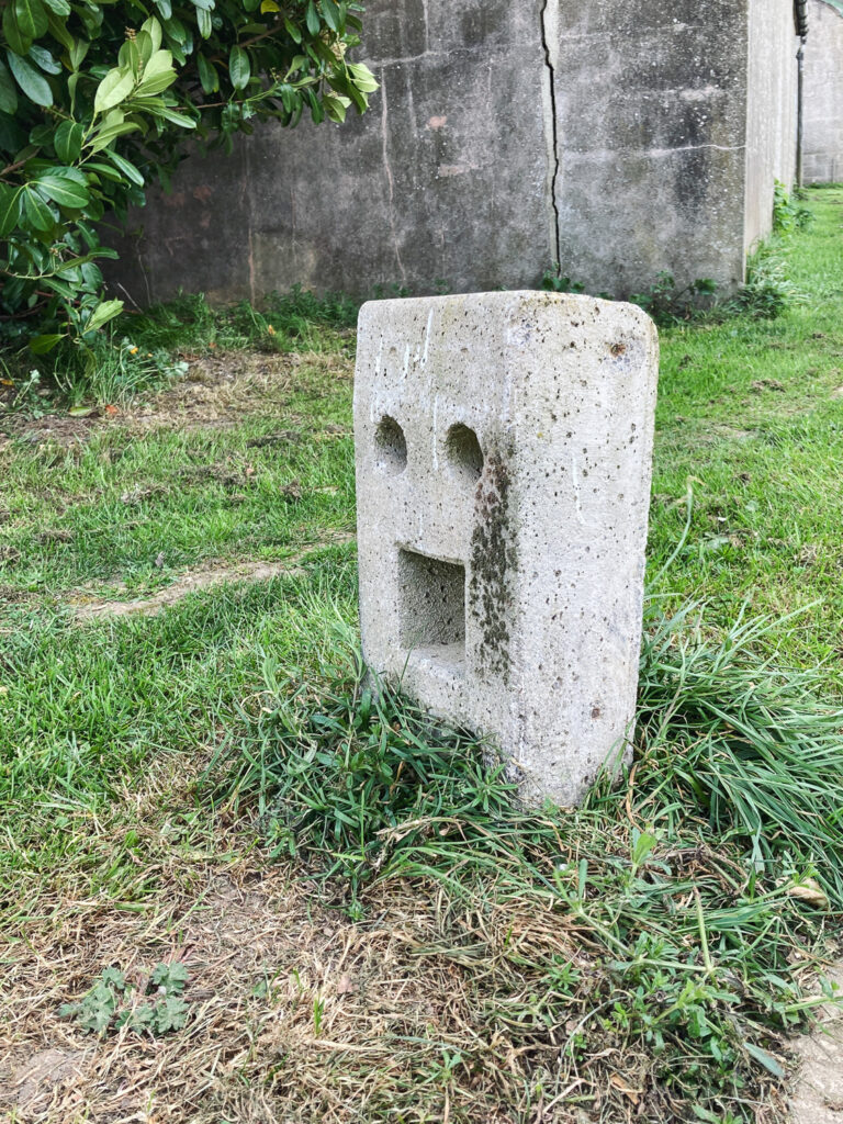 Photograph of a concrete bollard with holes that make it look like a face with a mouth wide open, with old buildings behind and grass around it.