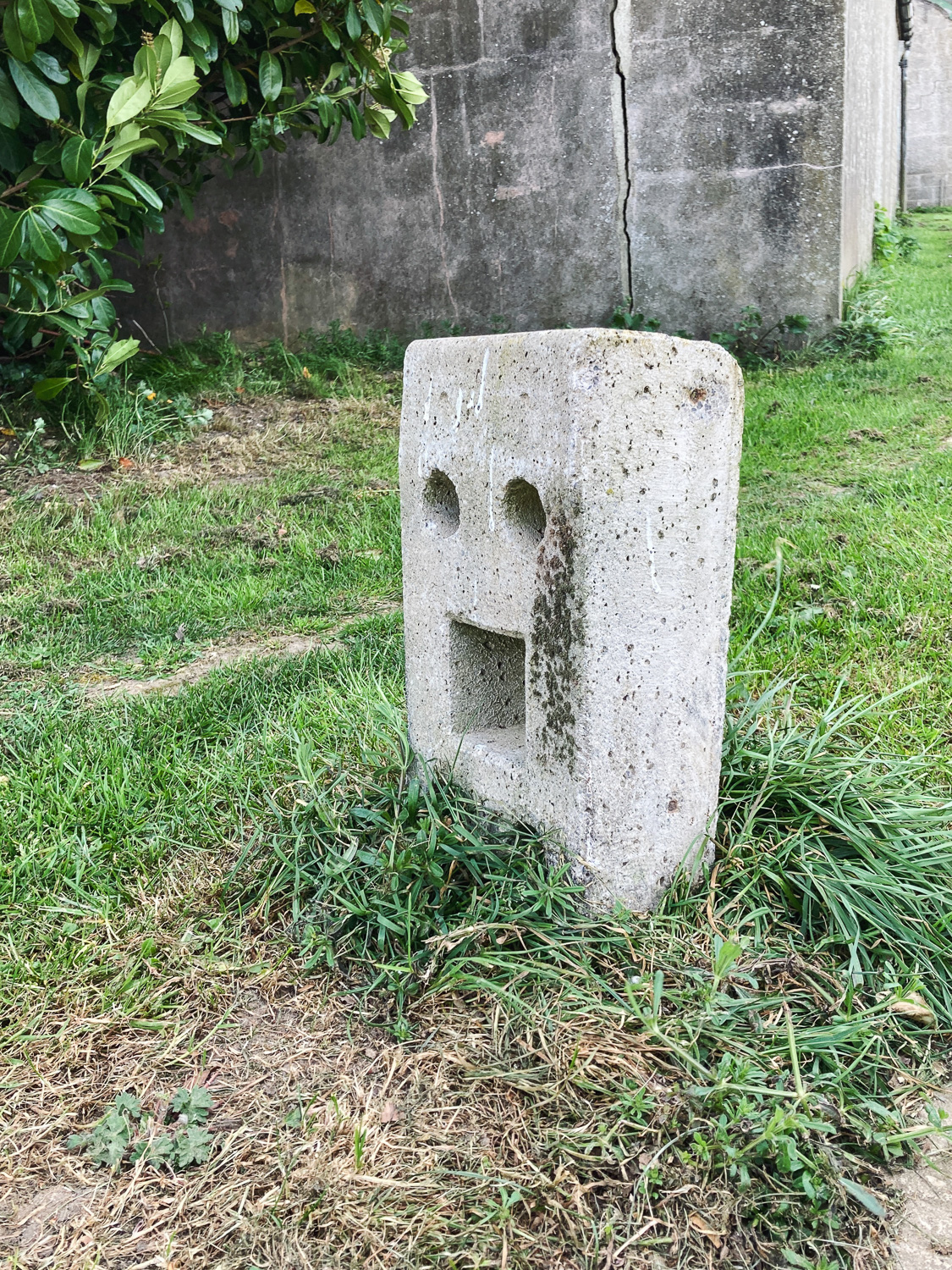 Hi! Photograph of a concrete bollard with holes that make it look like a face with a mouth wide open, with old buildings behind and grass around it.