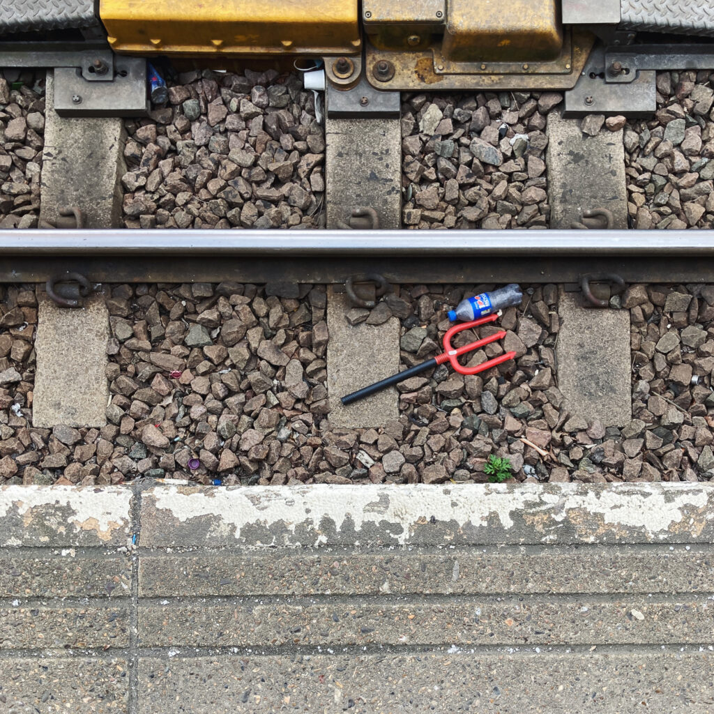 Photograph looking down at traintracks with a plastic devil's pitchfork in the gravel by the tracks, with the edge of the platform visible at the bottom of the frame.