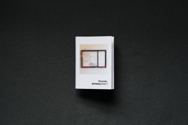 Photograph of a small foldy zine on a dark grey background. The cover shows an empty noticeboard with signs of wear, and text saying 'Obsolete. amasszine01'