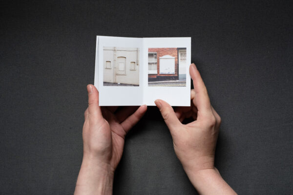 Photograph of hands holding a small foldy zine on a dark grey background. It is held open on pages with a photograph of a cream wall with 3 filled-in windows and an empty noticeboard to the left and a worn white empty noticeboard on a brick wall to the right.