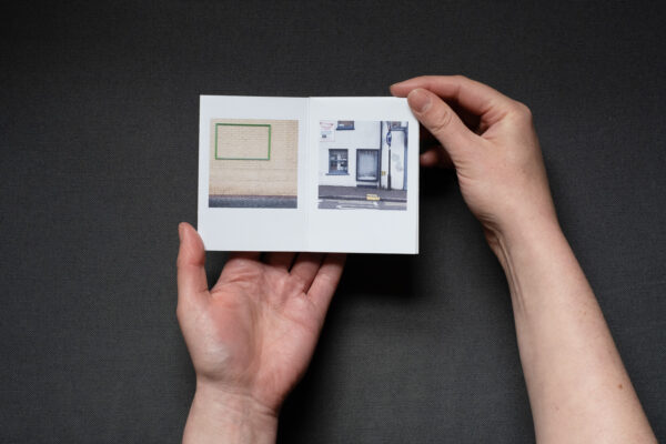 Photograph of hands holding a small foldy zine on a dark grey background. It is held open on pages with a photograph of an empty green frame on a brick wall to the left and a very worn blue frame next to a window and a streetlight at the side of a road to the right.