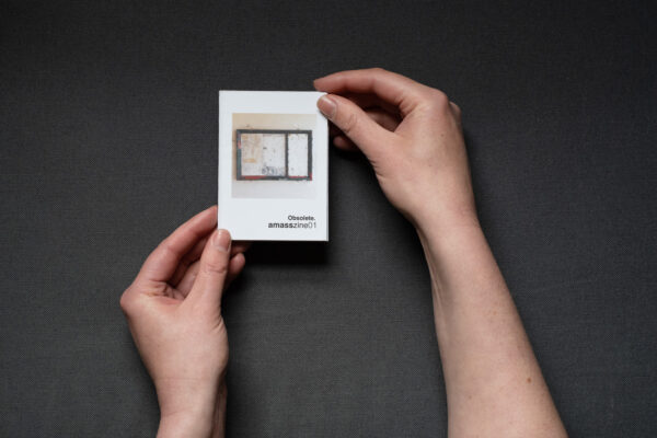 Photograph of hands holding a small foldy zine on a dark grey background. The cover shows an empty noticeboard with signs of wear, and text saying 'Obsolete. amasszine01'