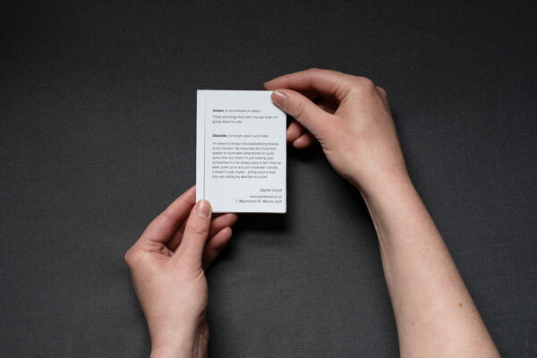 Photograph of hands holding a small foldy zine on a dark grey background. The back cover shows small text about the zine.