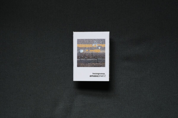 Photograph of a small foldy zine on a dark grey background. The cover shows a photograph of a smashed blue mug on double yellow lines against a curb, and text saying 'Incongruous. amasszine02'