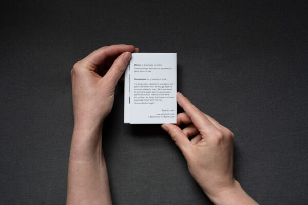 Photograph of hands holding a small foldy zine on a dark grey background. The back cover shows small text about the zine.