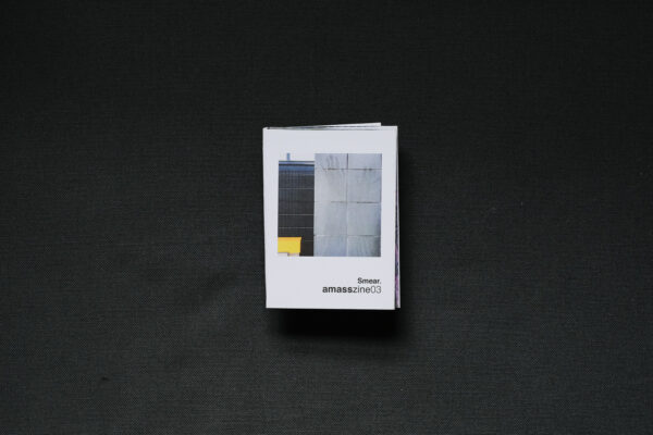 Photograph of a small foldy zine on a dark grey background. The cover shows a grey wall with greasy handprints smeared on it, with a darker wall and a yellow square slightly behind and to the left and text saying 'Smear. amasszine02'