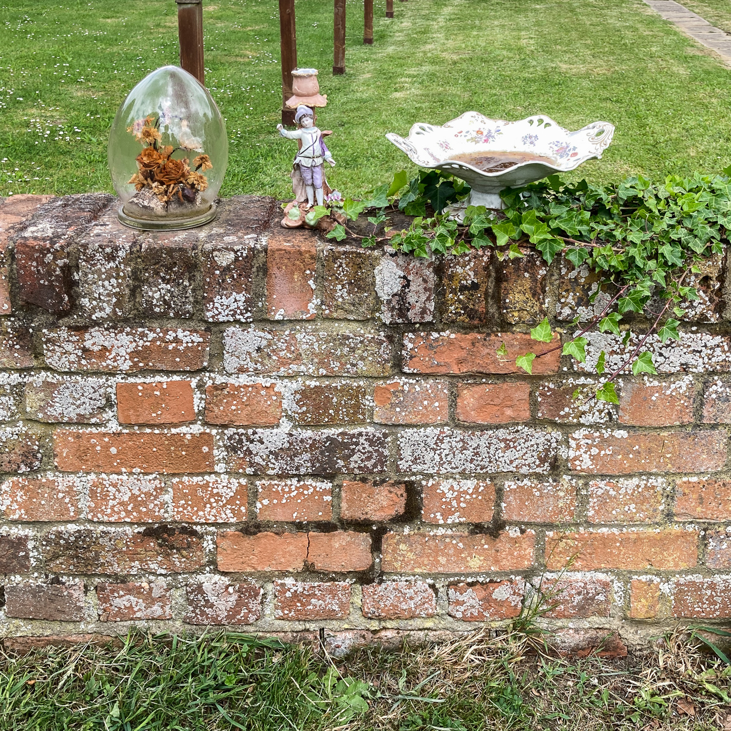 Curiosities. Photograph of a brick wall with a glass dome holding drived flowers, a statuette of a person and a decorative bowl with dirty water in it. Ivy is growing on the wall and lawn is visible behind and in front of the wall.