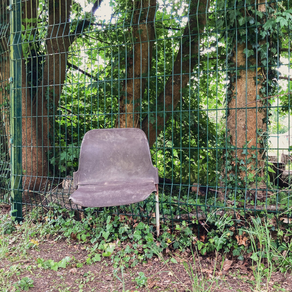 Photograph of a broken plastic school chair with only one leg, wedged into a wire fence, with trees behind.