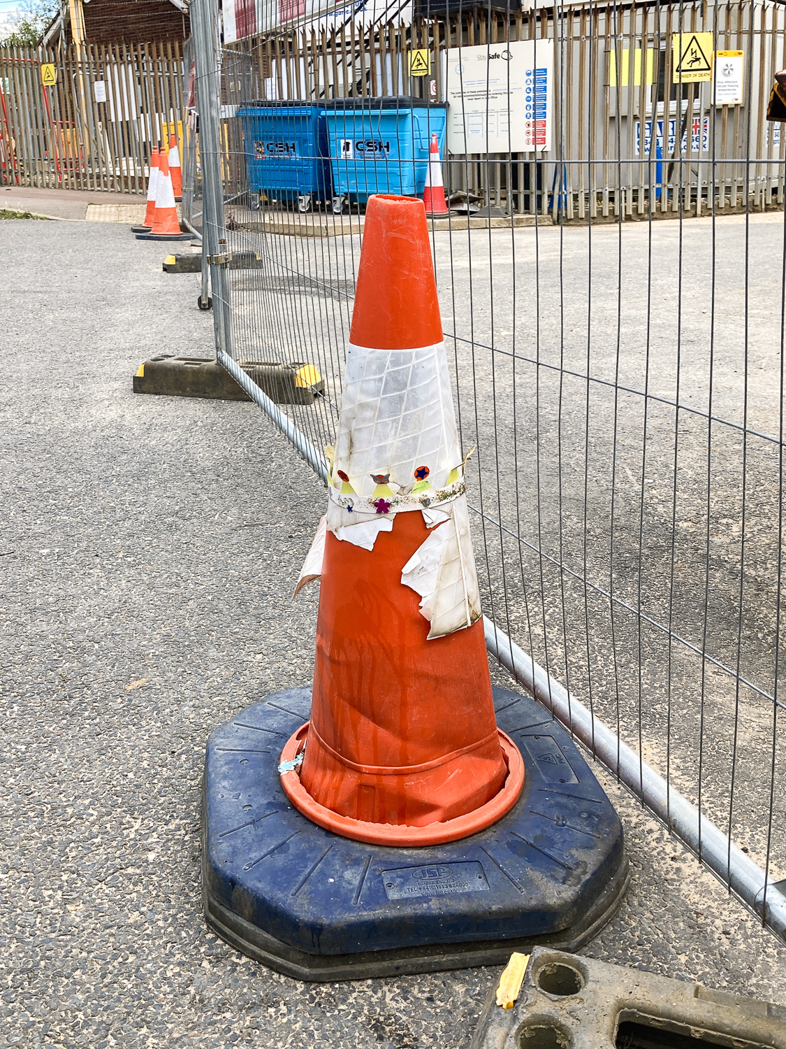 By Royal appointment. Photograph of a traffic cone with a child's handmade paper crown halfway down. Behind is a wire fence and bins at a construction site.