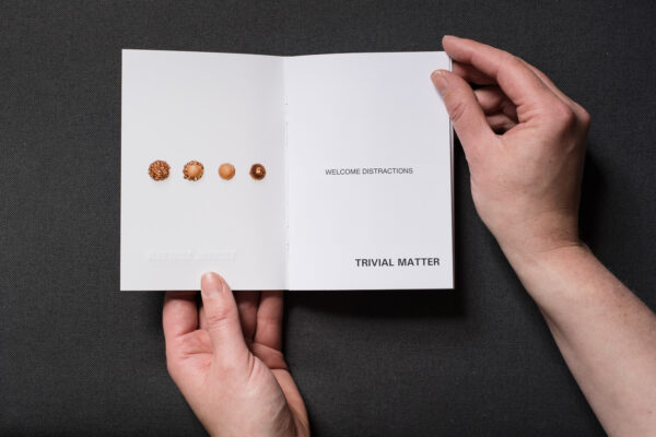 Photograph of TRIVIAL MATTER 01, a photo zine, held by hands on a dark grey fabric background. It is open on pages showing 4 stages of a hazelnut chocolate being eaten to the left and text to the right saying 'WELCOME DISTRACTIONS, TRIVIAL MATTER'.