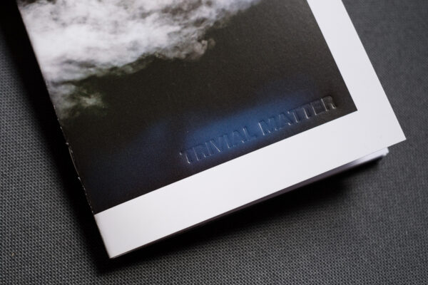 Photograph showing a detail of TRIVIAL MATTER 01, a photo zine, on a dark grey fabric background. 'TRIVIAL MATTER' is embossed with letter press.