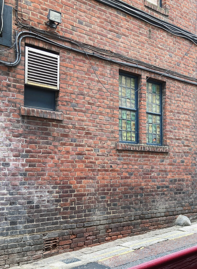 Photograph of a brick wall with cables along the top, an air vent to the left and a window with coloured glass that has been bricked up behind.