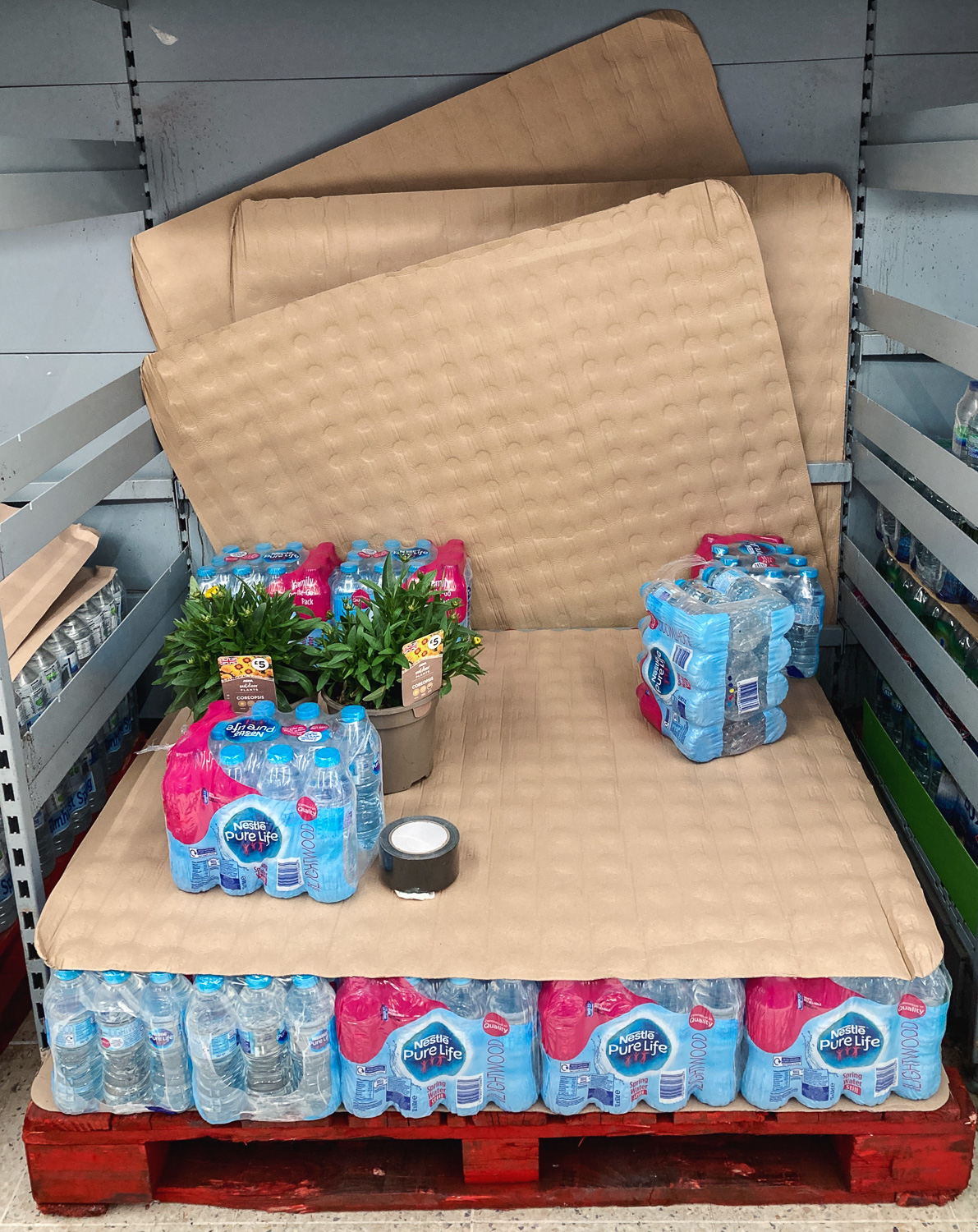 Change of plans. Photograph of a supermarket display of Nestle Pure Life water with potted plants and a roll of black tape that appear to have been put there.