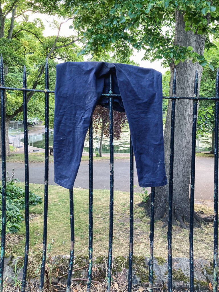 Photograph of a metal fence with park behind, with a navy pair of trousers hanging on the top.