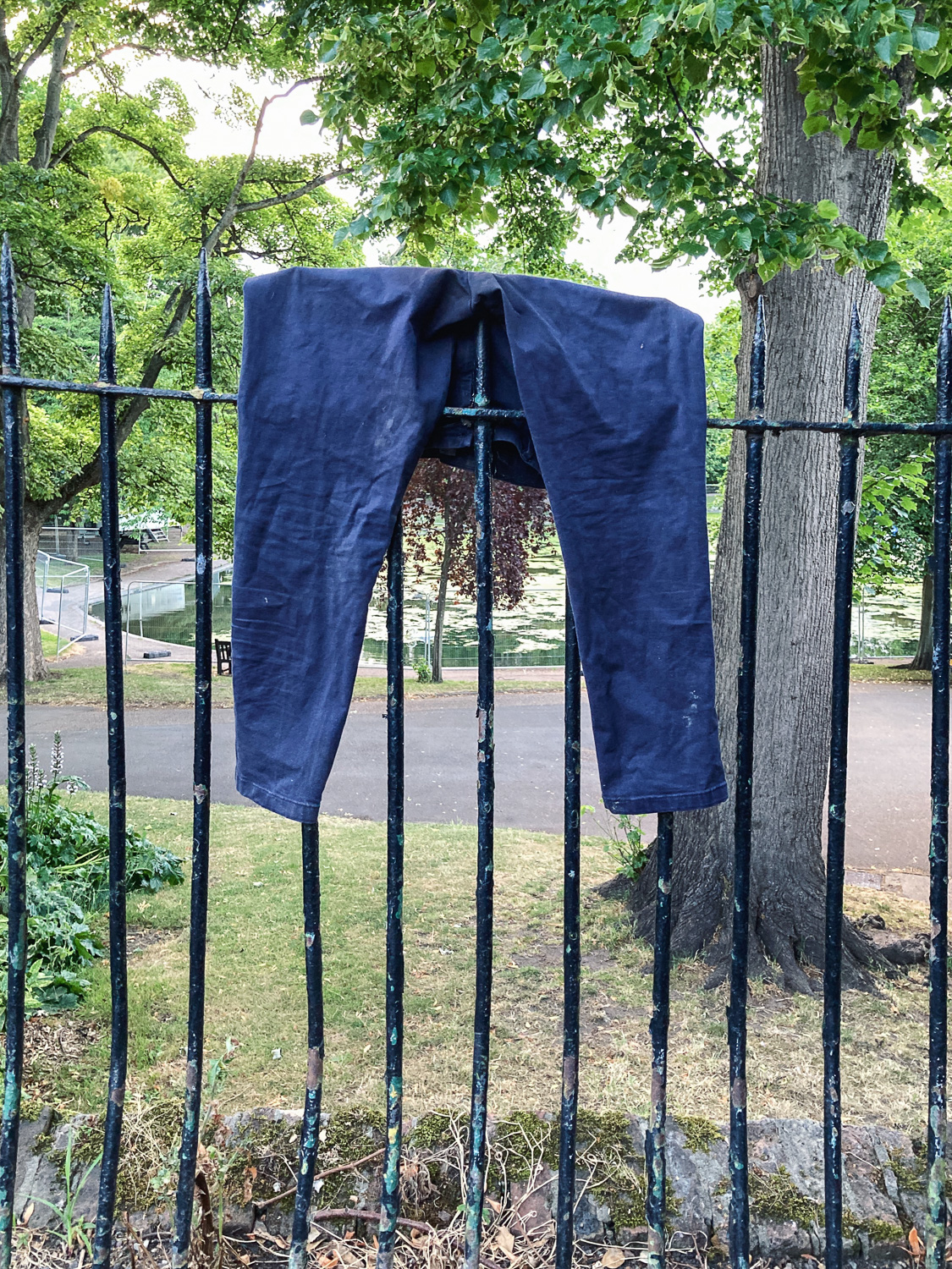 Hope they had a spare. Photograph of a metal fence with park behind, with a navy pair of trousers hanging on the top.