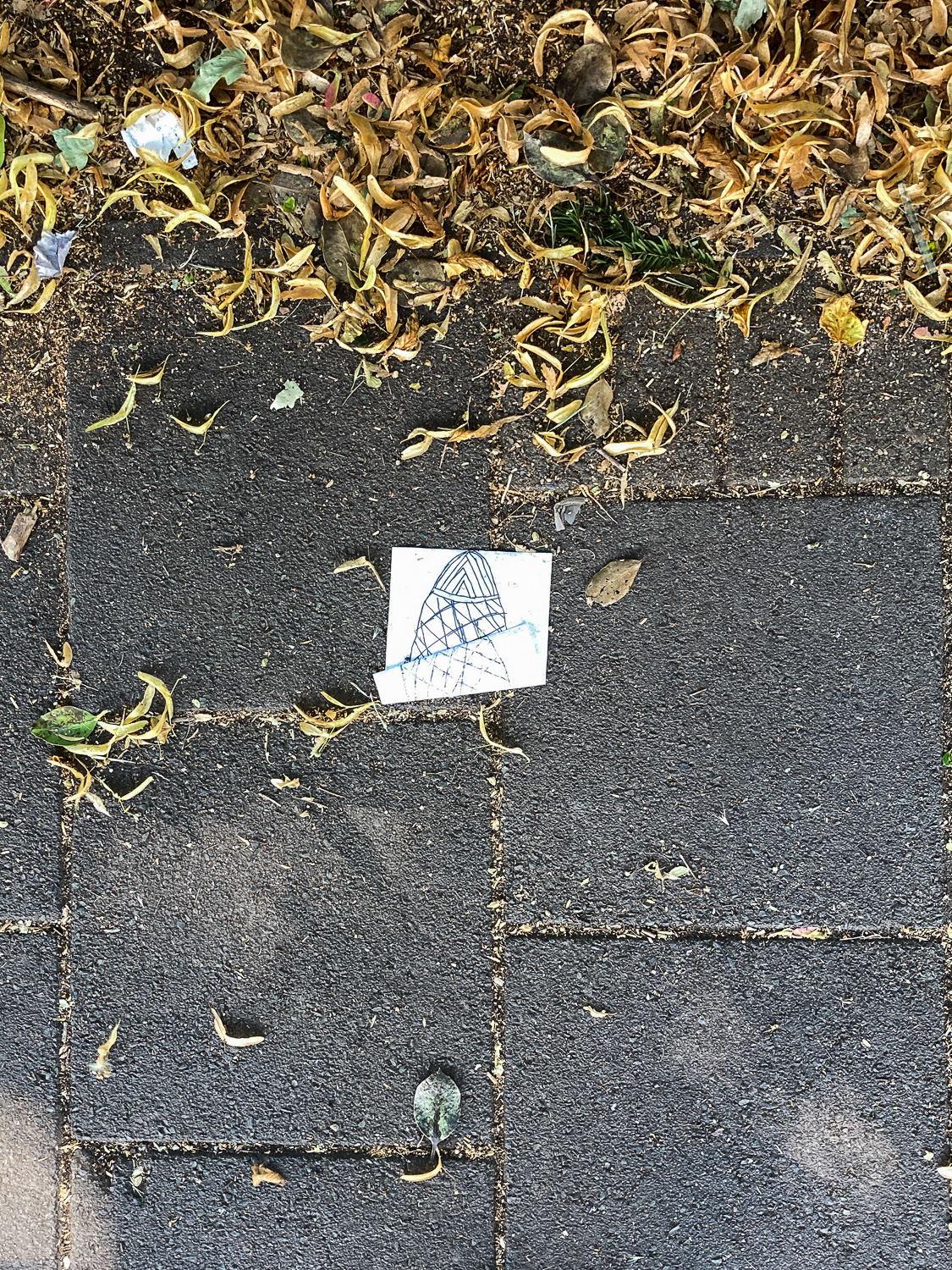 Glimpse of the Gherkin. Photograph of a pavement with a piece of paper that is folded but appears to show a pen drawing of the Gherkin. Dried leaves are to the top of the frame.