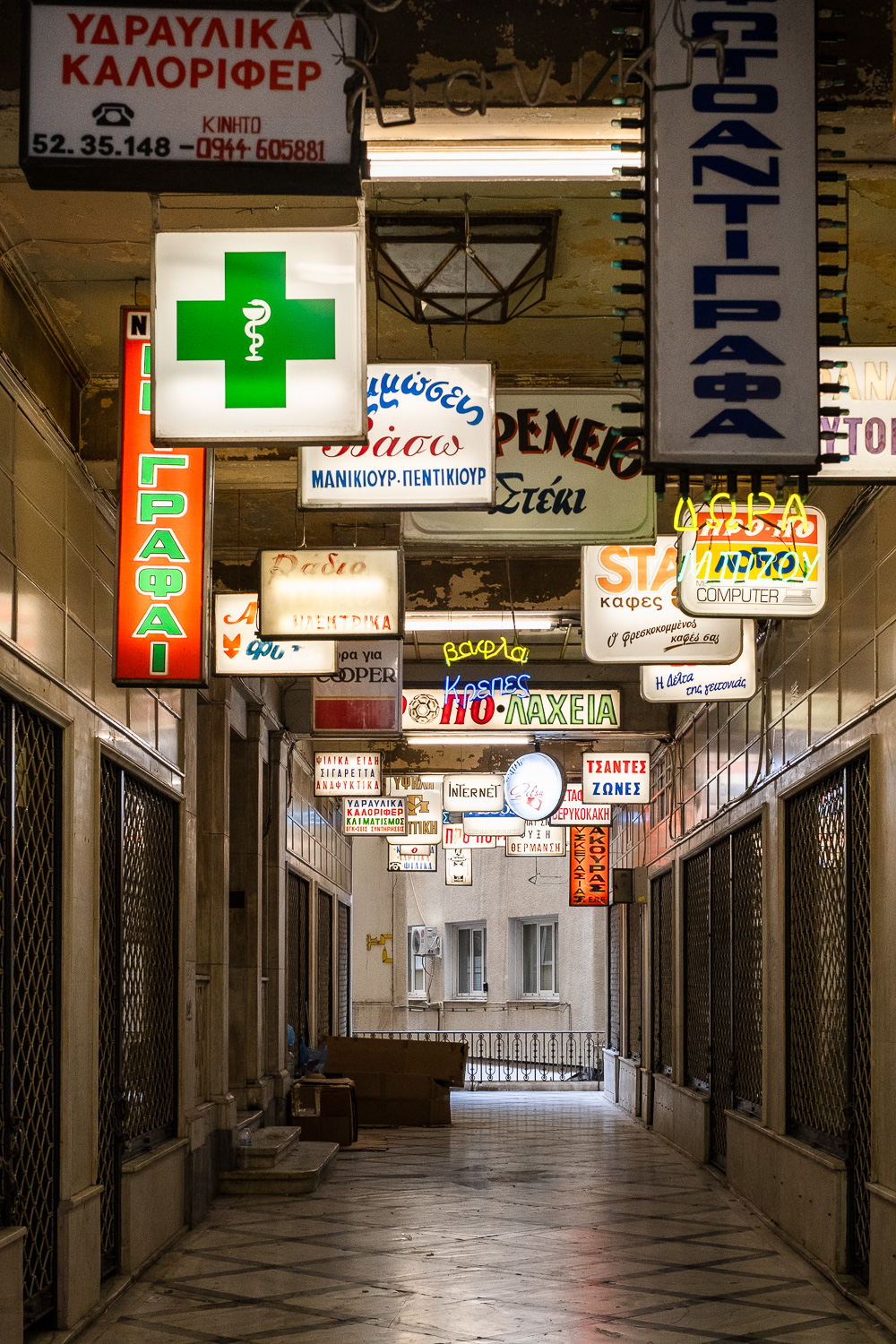 An alley I came back to photograph. Photograph looking into a walkway with shuttered up doors and windows and illuminated signs above with a pharmacy logo, and Greek text advertising shop names.