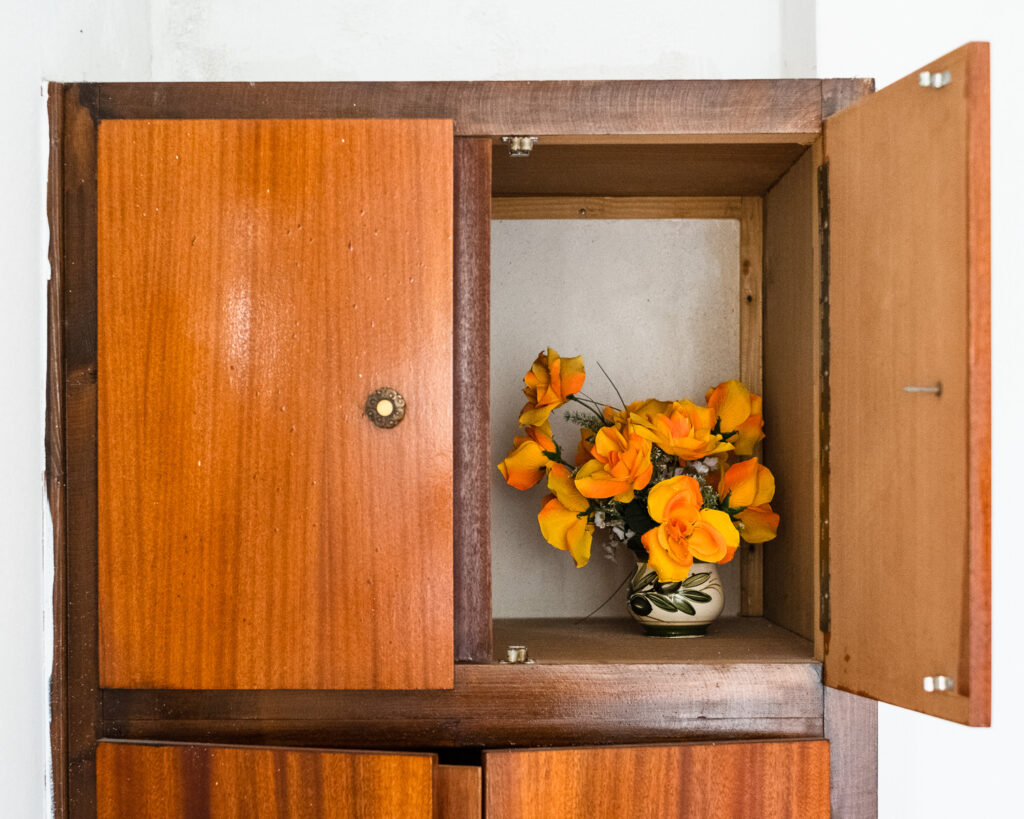 Photograph of a vase of artificial orange flowers in a wooden cupboard - right door open to show the flowers and left door closed, with an ornate handle.