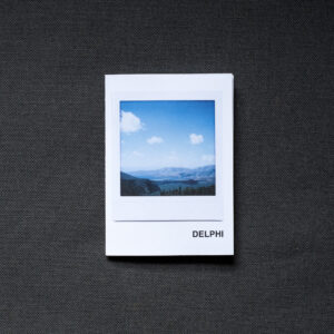 Photograph of an A7 zine on a dark grey fabric background. The zine has the word 'DELPHI' in the bottom right corner and an Instax Square print on the cover that shows a valley with mountains and blue sky with clouds above.