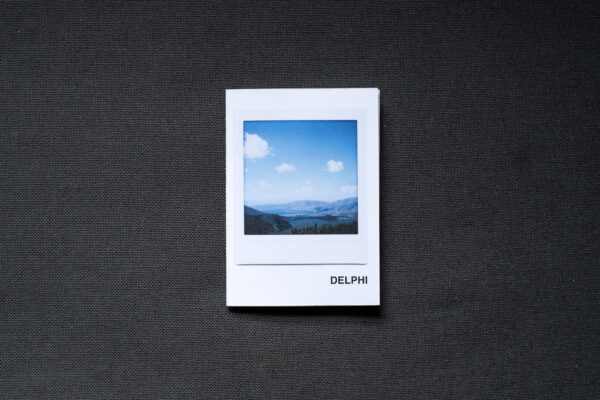 Photograph of an A7 zine on a dark grey fabric background. The zine has the word 'DELPHI' in the bottom right corner and an Instax Square print on the cover that shows a valley with mountains and blue sky with clouds above.