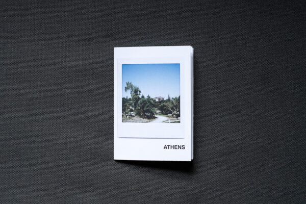 Photograph of an A7 zine on a dark grey fabric background. The zine has the word 'ATHENS' in the bottom right corner and an Instax Square print on the cover that shows a temple in the Ancient Agora with plants below and blue sky above.