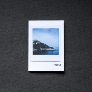 Photograph of an A7 zine on a dark grey fabric background. The zine has the word 'HYDRA' in the bottom right corner and an Instax Square print on the cover that shows the island of Hydra with sea in front and blue sky above.