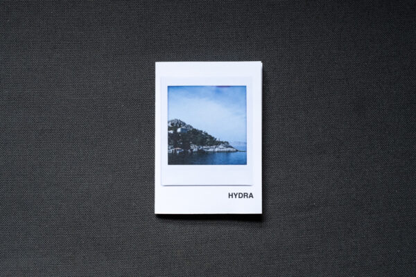 Photograph of an A7 zine on a dark grey fabric background. The zine has the word 'HYDRA' in the bottom right corner and an Instax Square print on the cover that shows the island of Hydra with sea in front and blue sky above.