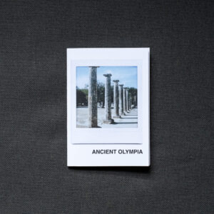 Photograph of an A7 zine on a dark grey fabric background. The zine has the word 'ANCIENT OLYMPIA' in the bottom right corner and an Instax Square print on the cover that shows columns in the Ancient Olympia archaeological site and blue sky above.